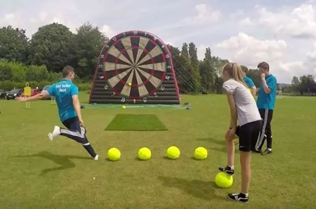 A player kicking a soccer ball towards a giant inflatable dartboard in a grassy field with others waiting for their turn