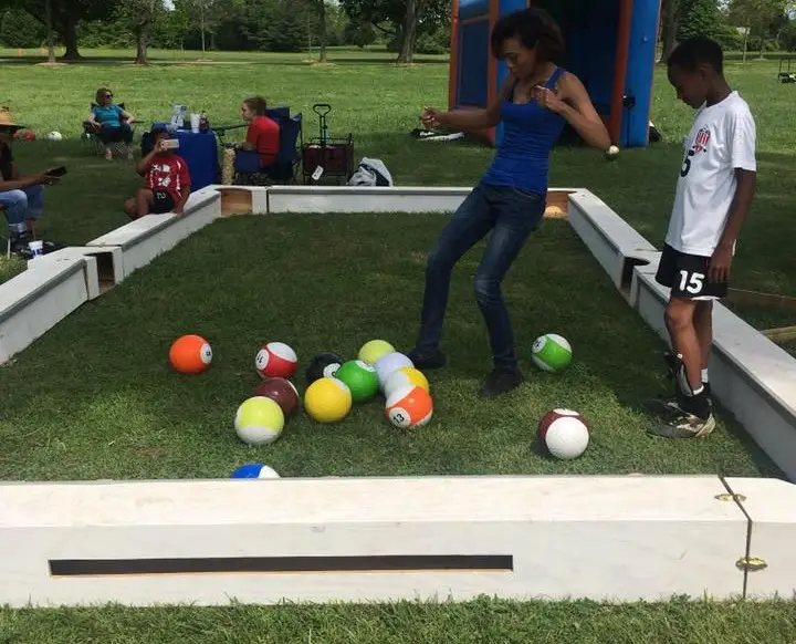 A woman and a young boy playing a giant billiards game with soccer balls designed as billiard balls in an outdoor setting