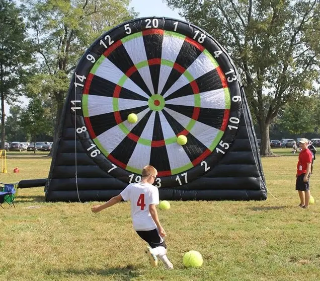 A player kicking a soccer ball towards a giant inflatable dartboard in a grassy field with others waiting for their turn