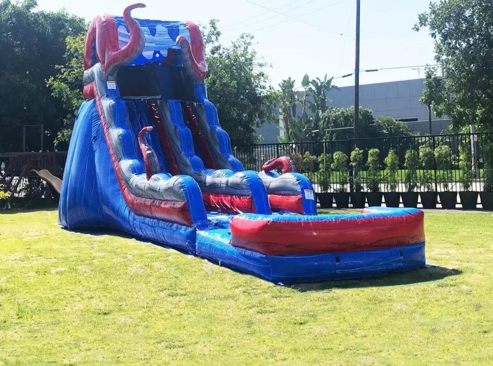 Inflatable water slide with blue and red colors featuring octopus tentacles design in a sunny outdoor setting with green grass and a fence in the background