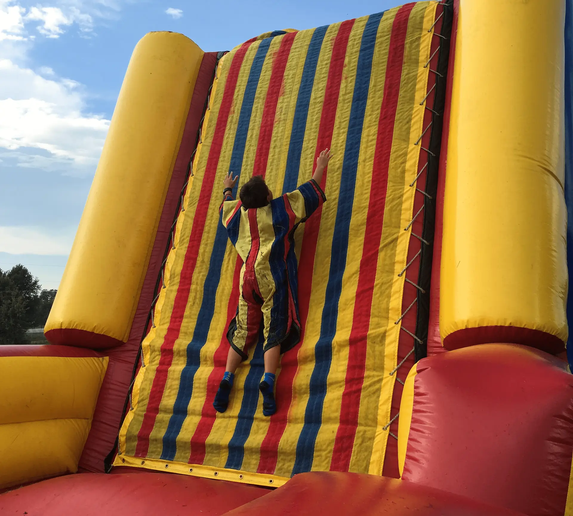 Child in a colorful striped outfit adhering to a Velcro climbing wall featuring bright yellow, red, and blue vertical stripes, attempting to climb higher on the inflatable structure